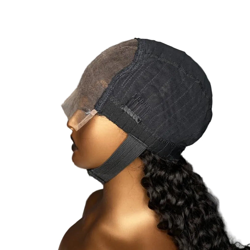 A dark charcoal gray lace front wig cap with a clear plastic protective cover over the forehead, shown on a mannequin head with long, dark curly hair. The mannequin is positioned in profile against a dark gray background with a white faux fur surface.